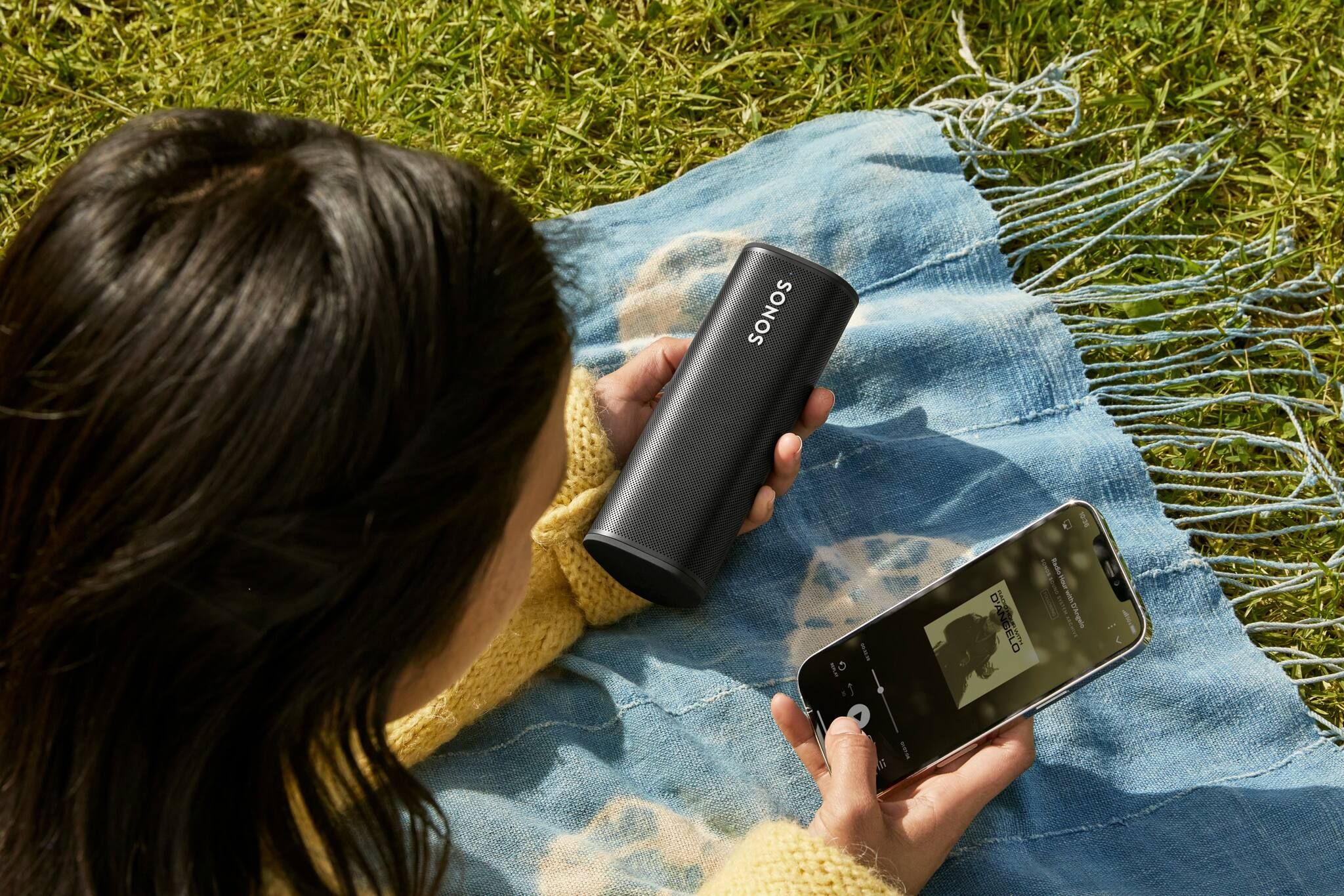 Lady lying on picnic mat, holding Sonos Roam and scrolling through music on phone