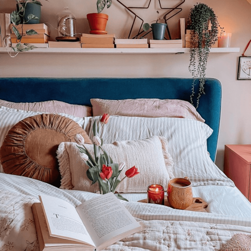a modern style bed with a blue velvet upholstered headboard, subtly striped bedding and textured decorative cushions. there are flowers, an open book and candles on the bed. by @my.interior.tales