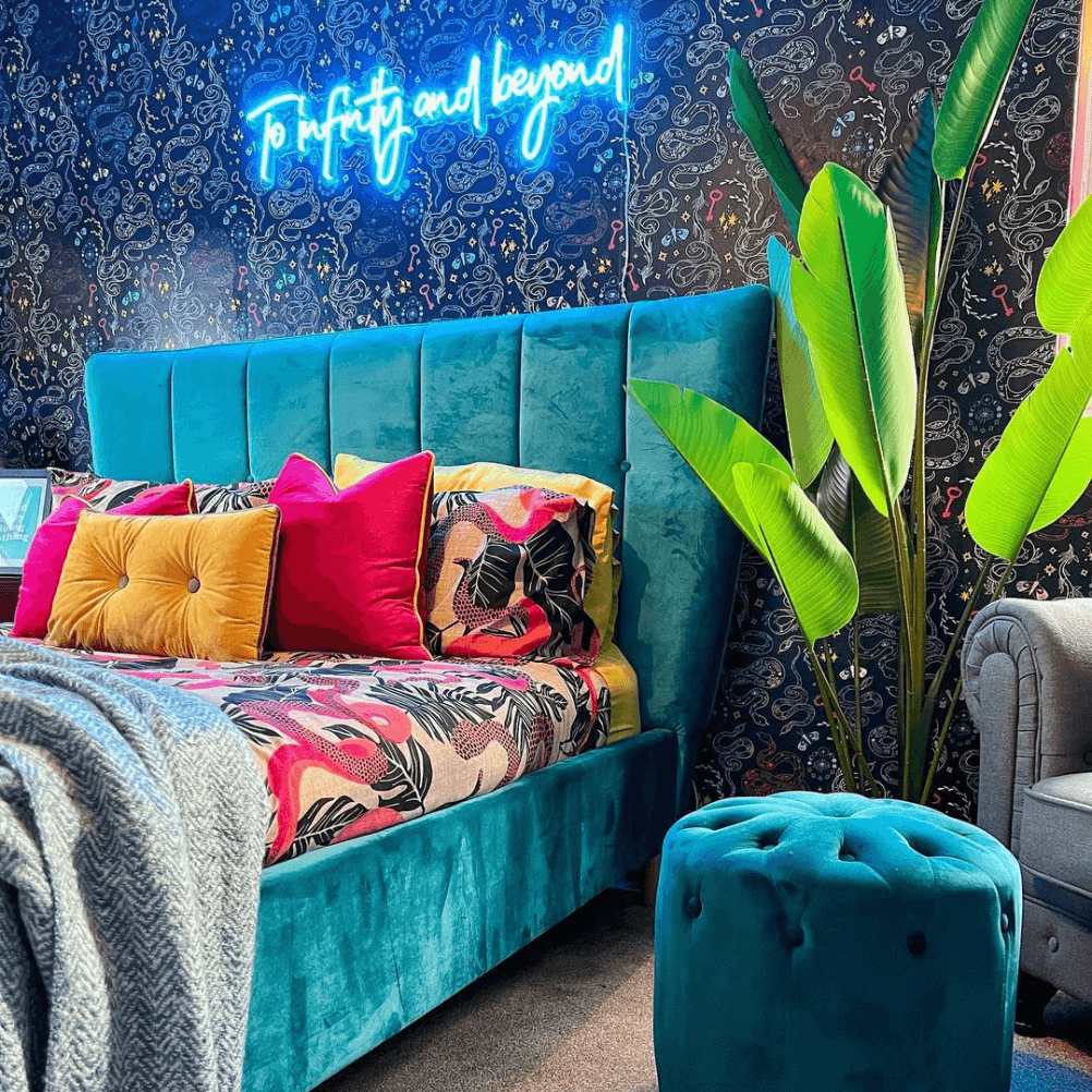 a bright and bold bedroom with dark patterned wallpaper, a neon light and a teal velvet bed. the bed is dressed in a pink snake pattern bedding. 