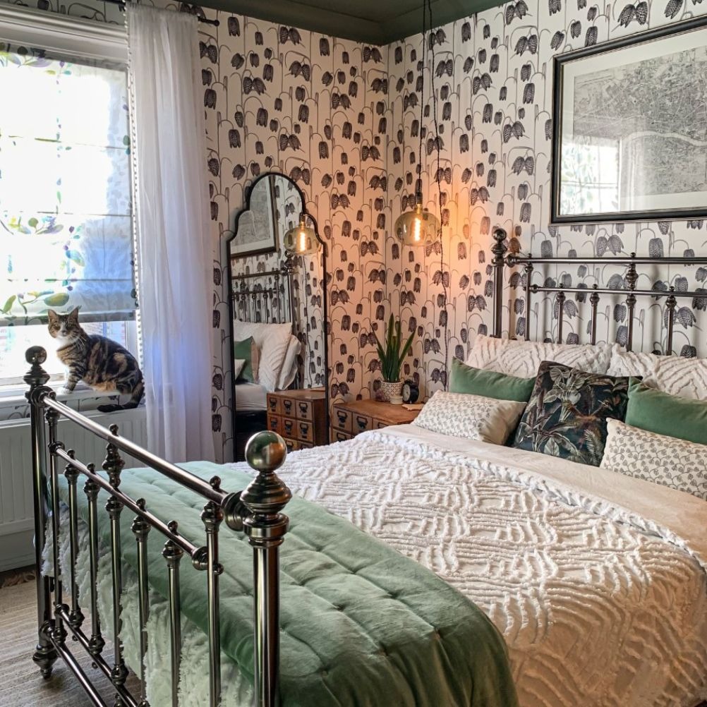 a victorian style room in cream and sage green. there's a black and white floral patterned wallpaper on the wall, a metal framed bed, textured bedding and a cat on the window sill. by @stove_pipe_house