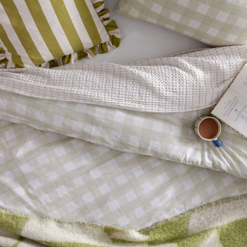 A cosy bedroom scene with green and white checkered pillows, a warm blanket, and a coffee cup next to a book. 