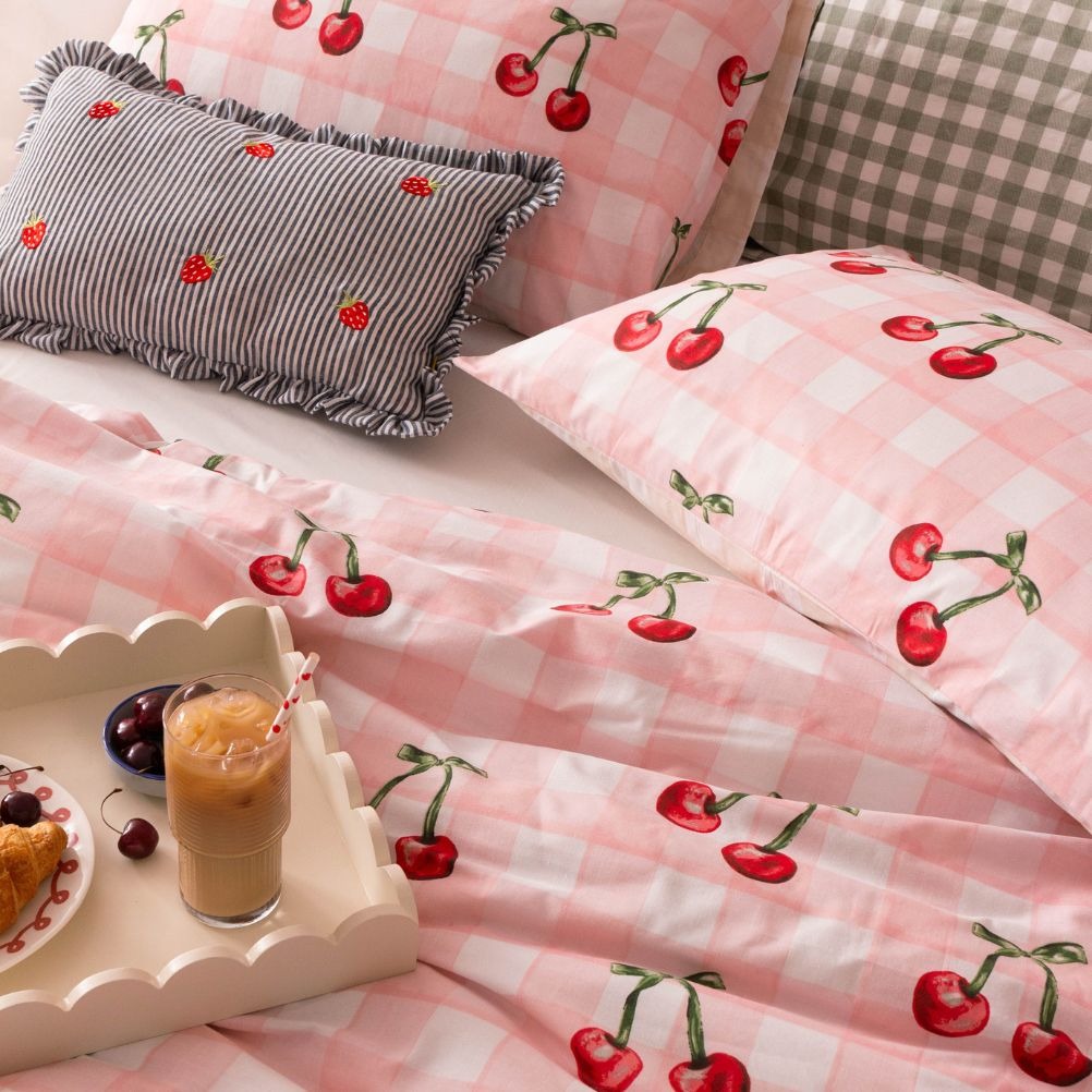 A bed with cherry-patterned pink check bedding, a striped, strawberry embroidered cushion, and a tray with iced coffee, fresh cherries and pastries.