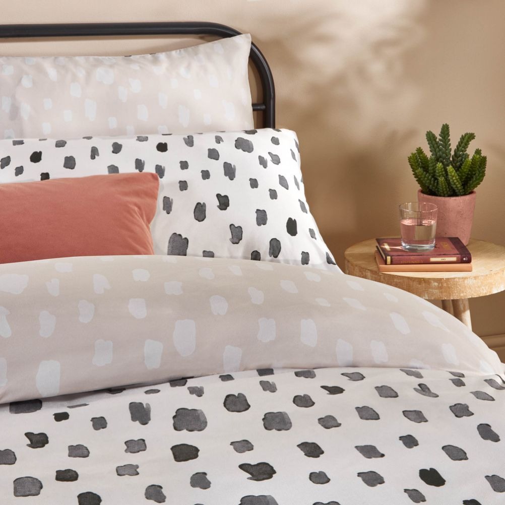 A neatly made bed with black and gray spotted bedding, a pink cushion, and a bedside table with plants and books.