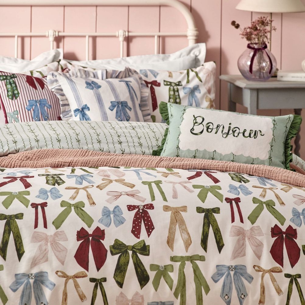 A bed with colourful bedding featuring various ribbon patterns and a cushion that says 'Bonjour'. 