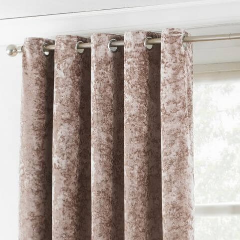 A crushed velvet curtain panel in a pale oyster pink shade, hung on a silver curtain pole in front of a window and a white wall.