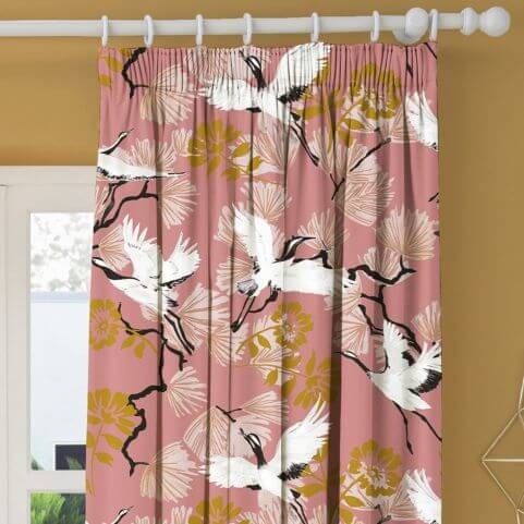 A pink curtain panel with a printed floral and bird design, hung on a white curtain pole in front of a gold background.