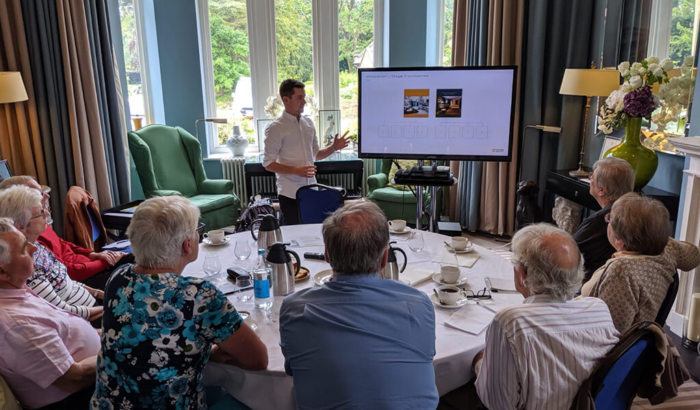 A member of the Motionspot team stood next to an interactive screen showing visual imagery to 8 older people sat around a table who all appear to be engaged.