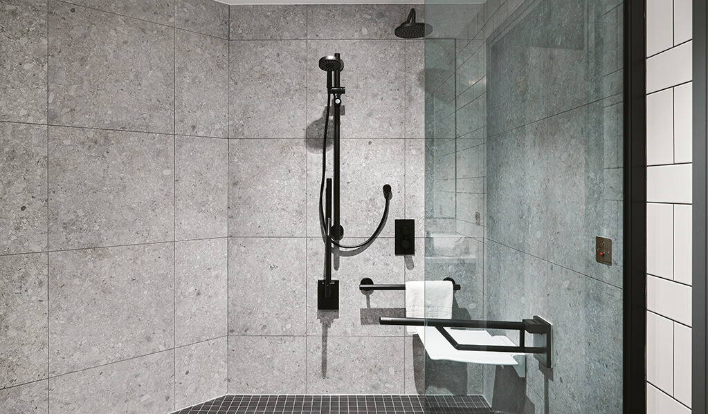 Hotel Brooklyn accessible shower area featuring large grey quartz tiles with matt black Doc M pack, folded shower seat and tinted shower glass screen.
