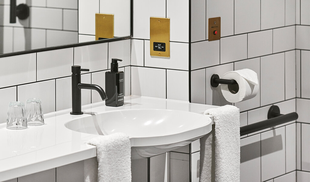 Hotel Brooklyn white tiled, black tile trim bathroom details, showcasing the integrated hand grip basin, matt black brassware and brass plated accessories. 