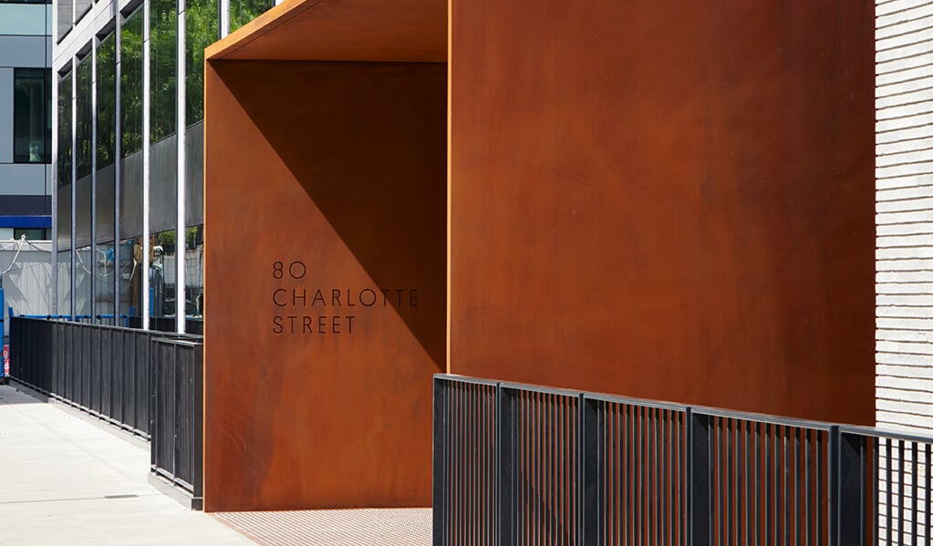 Copper coloured exterior of entrance view reading 80 charlotte street, attaching to the front of grey brick building tower. 
