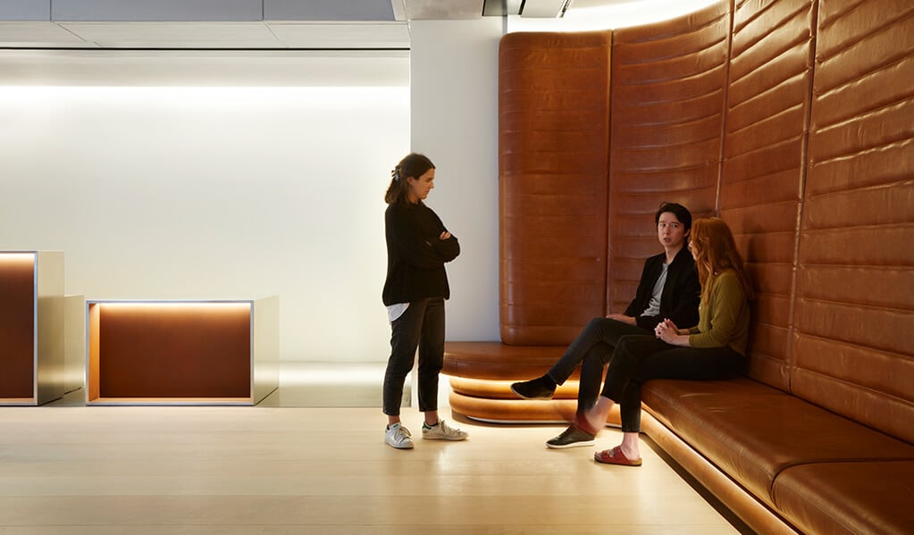 Tall backed copper coloured leather seating with lighting underneath, curving around interior walls, with people seated and standing in discussion. 
