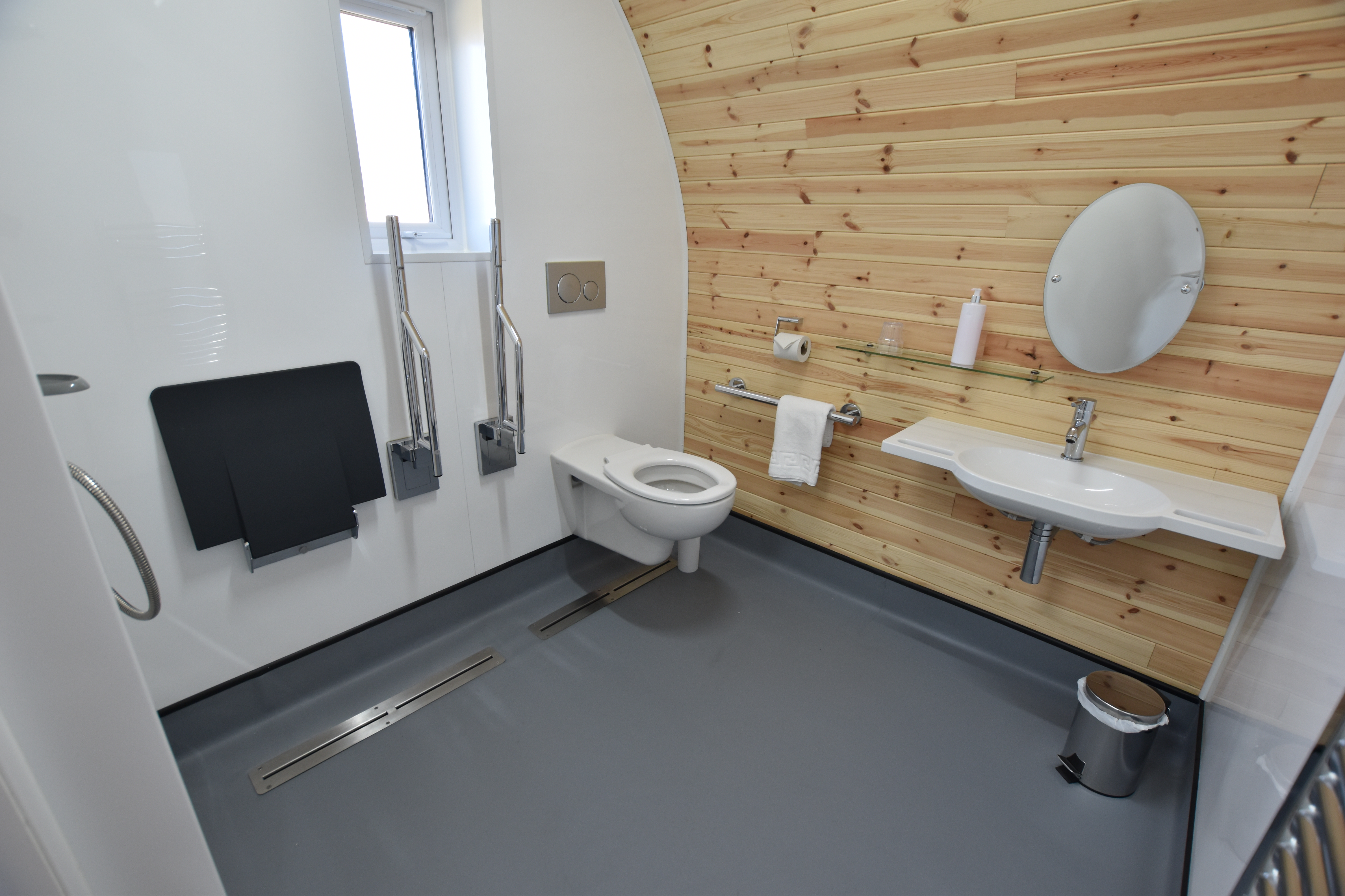 Accessible OmniPod bathroom featuring curved natural wood clad wall, wall-mounted basin with integrated hand-grips, toilet and shower seat with grab rails. 