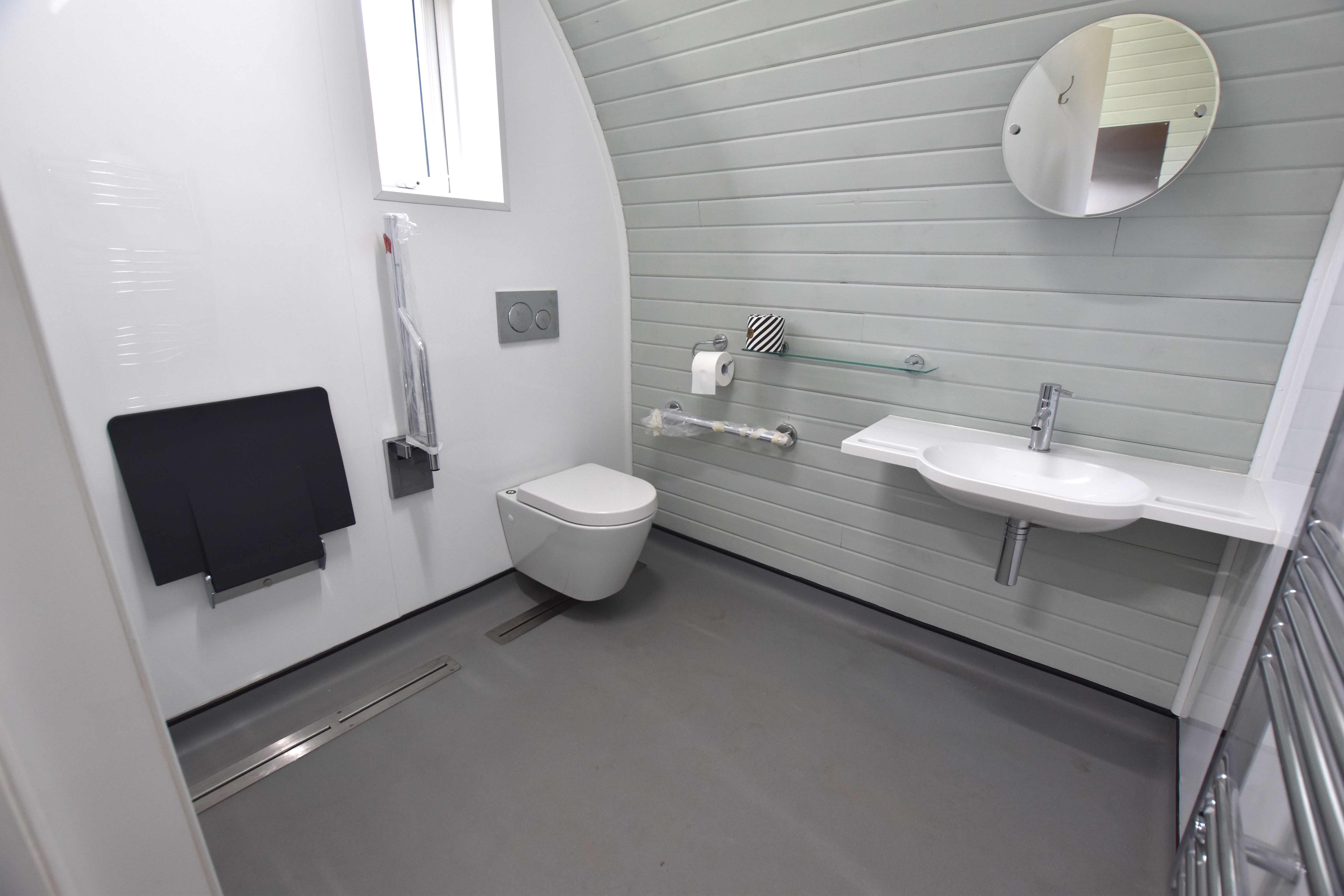 Accessible OmniPod bathroom featuring curved grey painted wood clad wall, wall-mounted basin with integrated handgrips, toilet, and shower seat with grab rails.