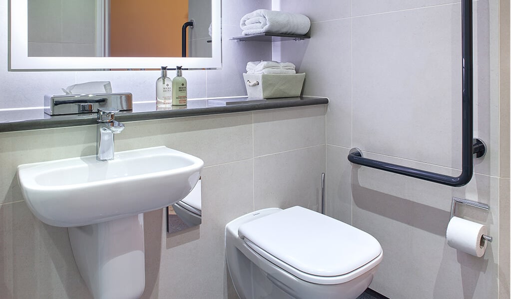 Bathroom with white wall hung toilet, L shaped hand rail and illuminated mirror above white hand basin with lovely chrome finishes. 
