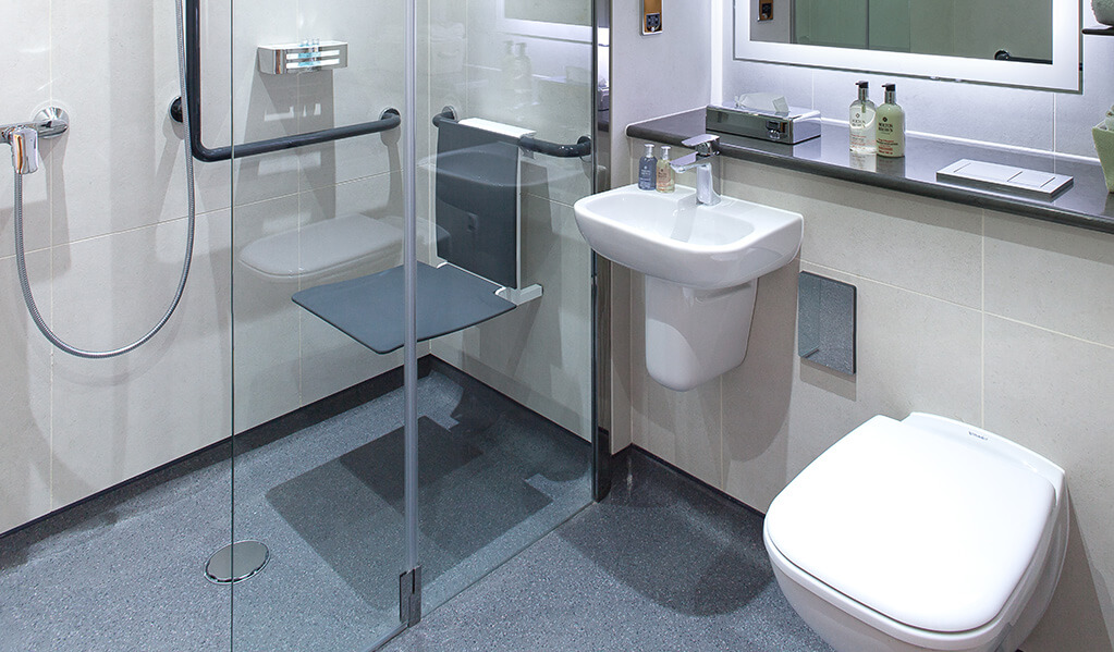 Bathroom with wall hung white toilet, small white basin and a bifolding shower screen which homes a wall hung charcoal coloured shower seat and L shaped shower rail. 
