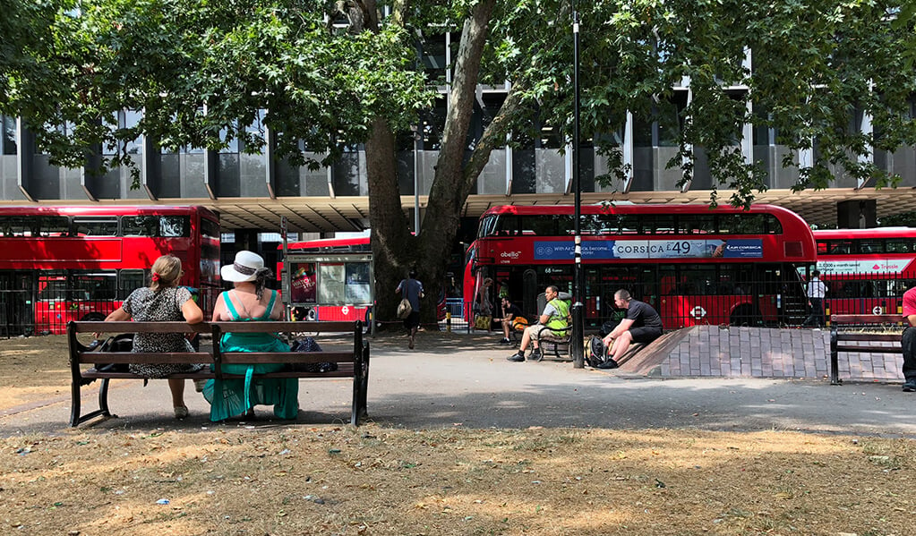 Exterior image of a red London double decker bus with people sat on benches admiring them. 
