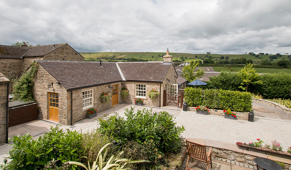 Exterior large angled view of the cottage in the Dales, L shaped building with large windows and light wood doors with a view of the garden which has lots of plants and tidy drive.  
