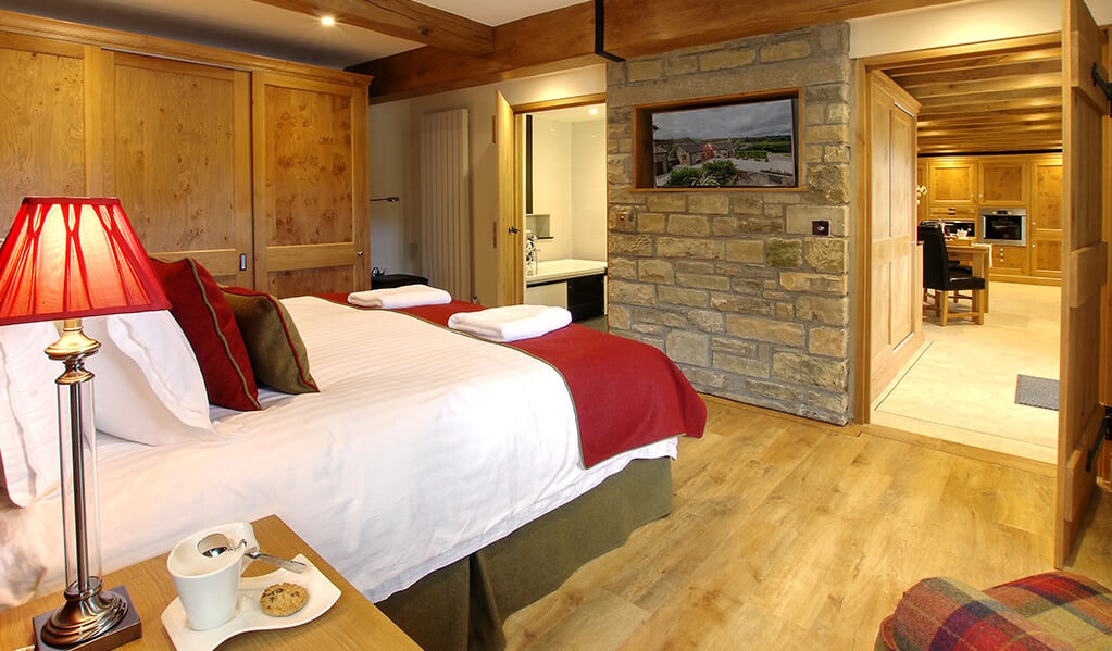 Light wood interior bedroom with exposed brick that holds an integrated television, double bed in the centre of the back wall with a view to the bathroom and kitchen.  
