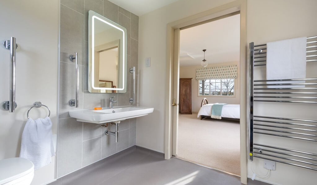 Neutral tone bathroom with chrome detailing throughout with illuminated mirror with view of cream coloured bedroom with lovely teal blue soft furnishings. 
