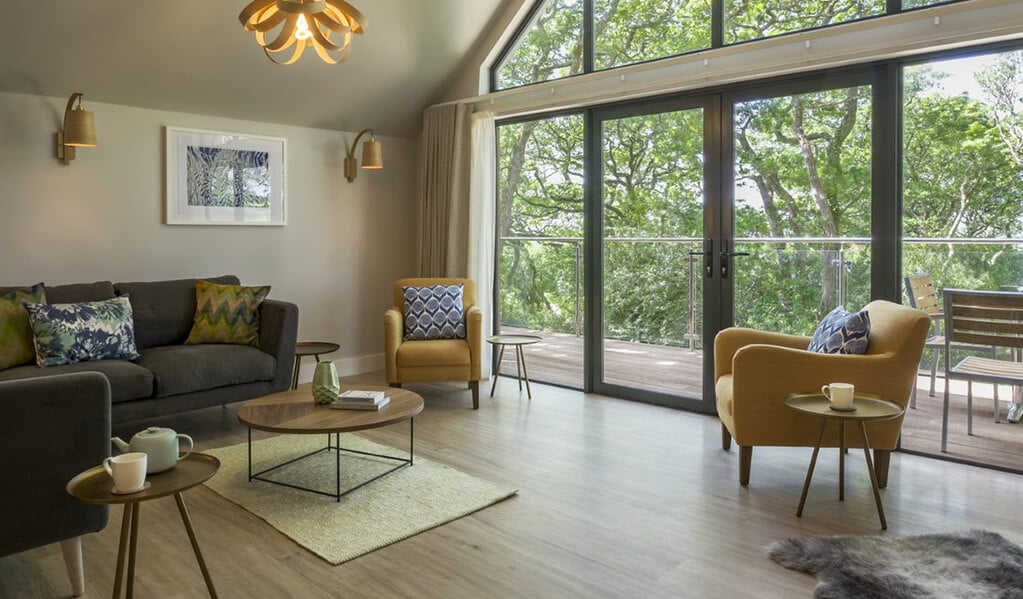 Open plan view of living area, mustard and grey coloured furnishings and brown wood flooring. Gorgeous floor to ceiling windows and double doors with view onto the balcony, surrounded by lovely bright green trees.
