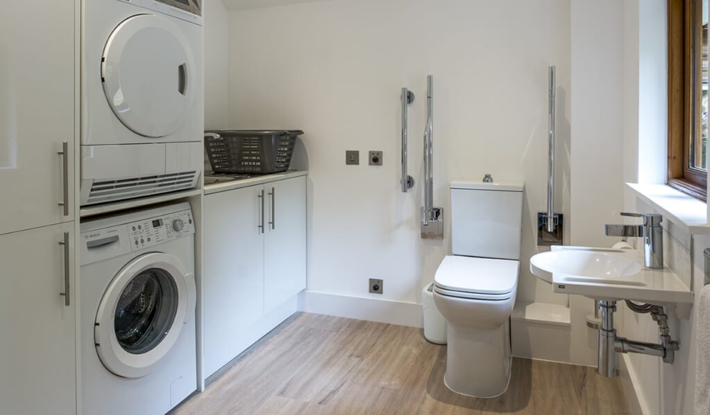 Utility room with white washing machine and dryer, an accessible modern toilet with chrome finished grabrails. 
