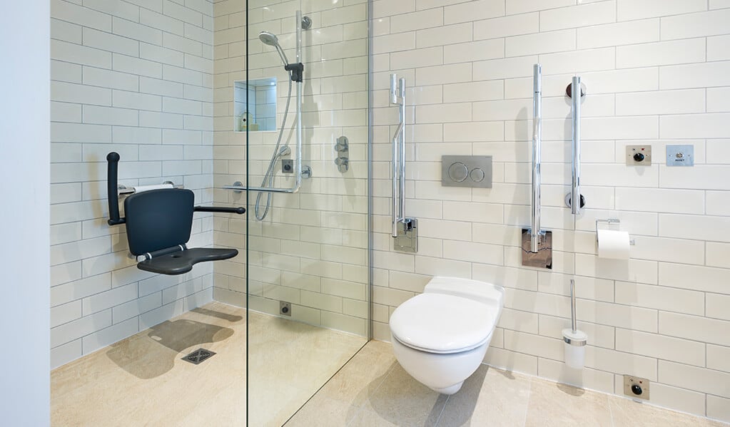 White modern tiled accessible bathroom with chrome finishes and large open shower with a matt black wall hung shower seat.
