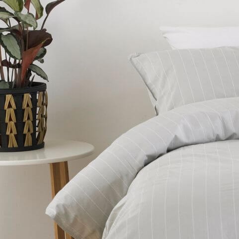 A grey 100% cotton duvet cover set with a white vertical pinstripe design, made on a bed in a neutral room next to a white side table holding a potted plant.