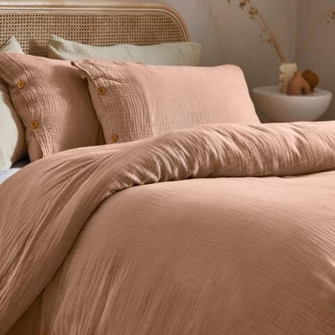 A 100% cotton muslin duvet cover set in an earthy pink clay shade, made on a bed with coordinating neutral cushions in a neutral room. 