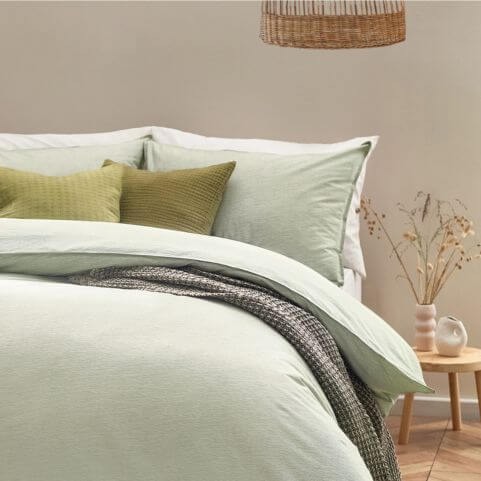 A 100% cotton bedding set in a khaki green shade with a subtle striped design, made on a bed with coordinating green scatter cushions and a grey throw.