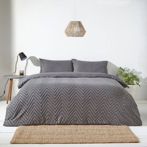 A charcoal grey 100% cotton duvet cover set with a tufted geometric design, presented in a neutral bedroom along with a side table, table lamp, a woven rug and an indoor plant.