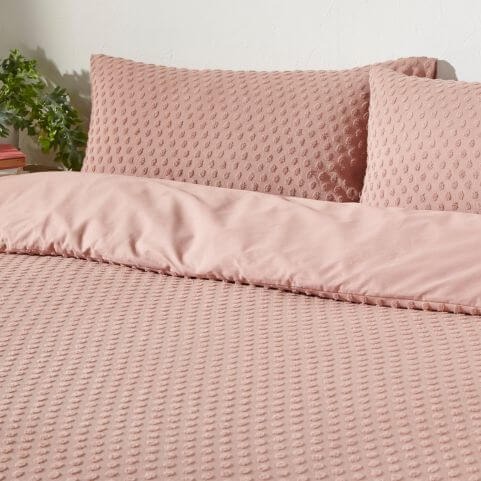 A blush pink 100% cotton duvet cover set with a tufted polka dot design, presented in a white-walled bedroom next to a hide table holding an indoor plant.