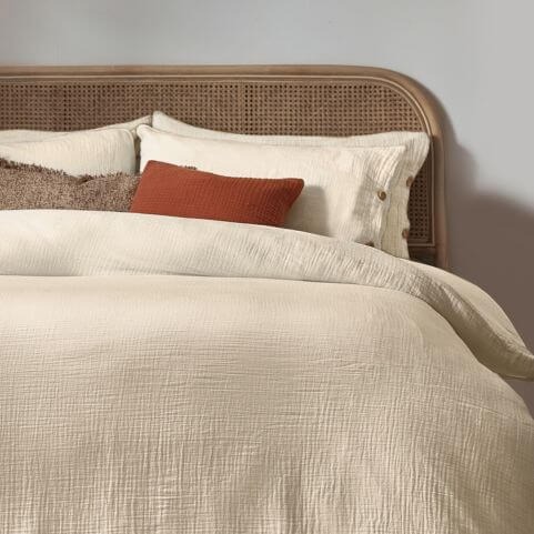 A cotton muslin duvet cover set in a natural off-white shade, presented on a bed with a wooden bedframe alongside coordinating scatter cushions.