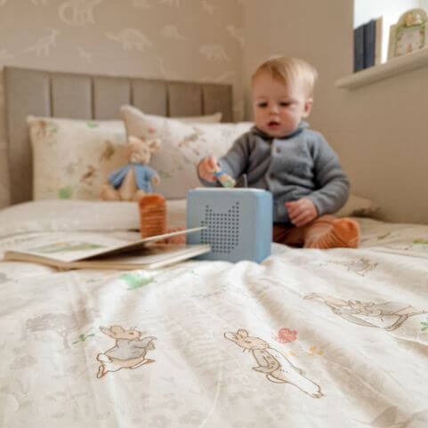 A natural beige coloured 100% cotton duvet set with a signature Peter Rabbit design, presented on a bed with a child and several toys.