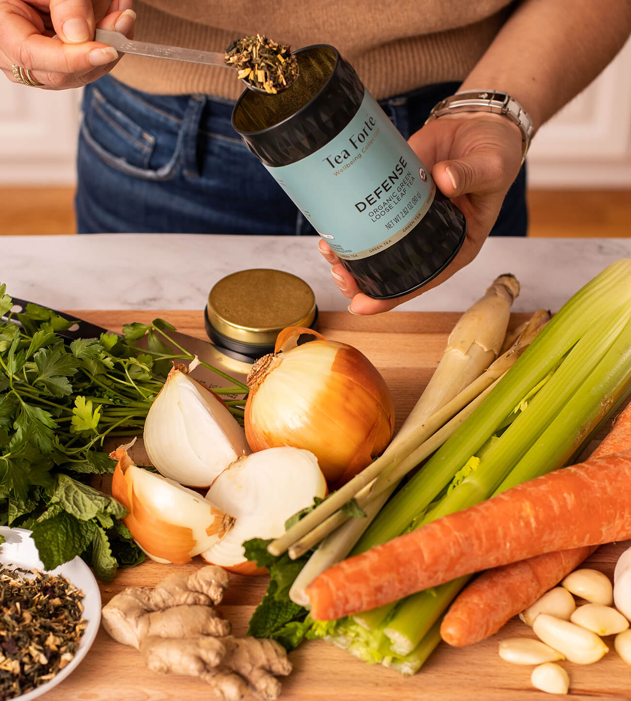 Scooping tea from a Defense Loose tea canister while standing over a large assortment of vegetables in a kitchen