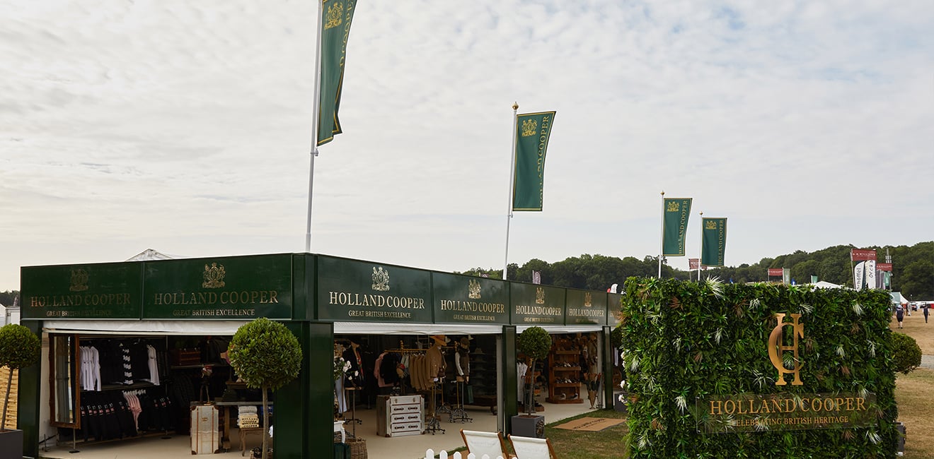 Wide angle image of Holland Cooper event stand at Game Fair 2022. Green flags and branding with a large flower wall with gold HC Holland Cooper writing on it.
