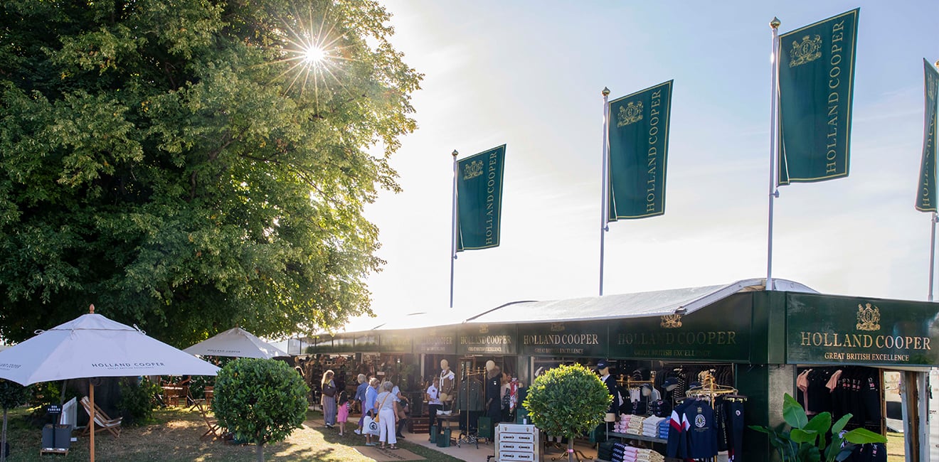 Wide angle image of Holland Cooper event stand at Burghley Horse Trials 2022. Green flags and branding with sun shining through a green tree onto the stand.