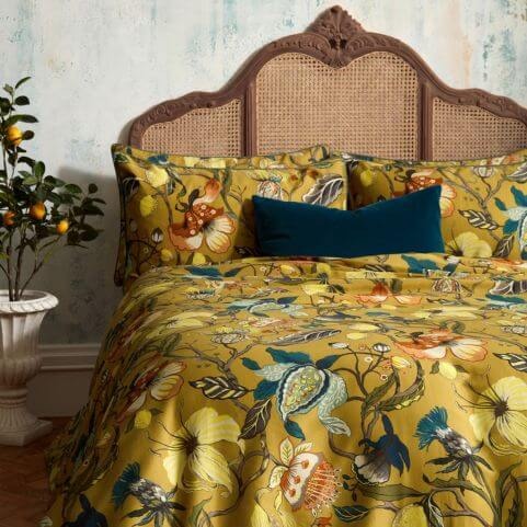 A luxury sateen ochre duvet cover set with a vibrant floral design, made on a bed with a traditional wooden headboard, a teal cushion and watercolour blue walls, with a small orange tree in a plant pot to the left hand side.