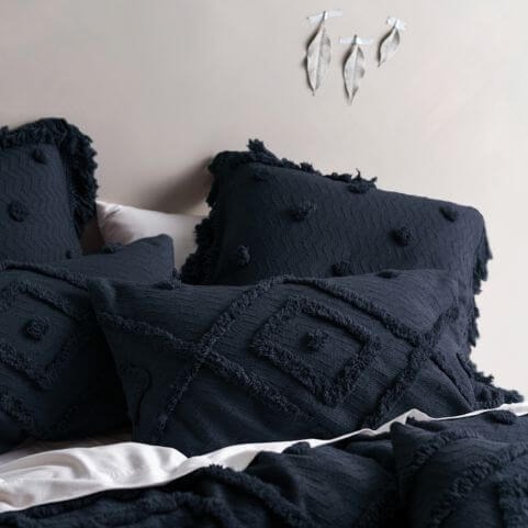 A selection indigo-coloured shams and pillowcases with a tufted geometric and polka dot tufted designs, arranged loosely on white bedding in front of a grey wall decorated with three leaves stuck to it.
