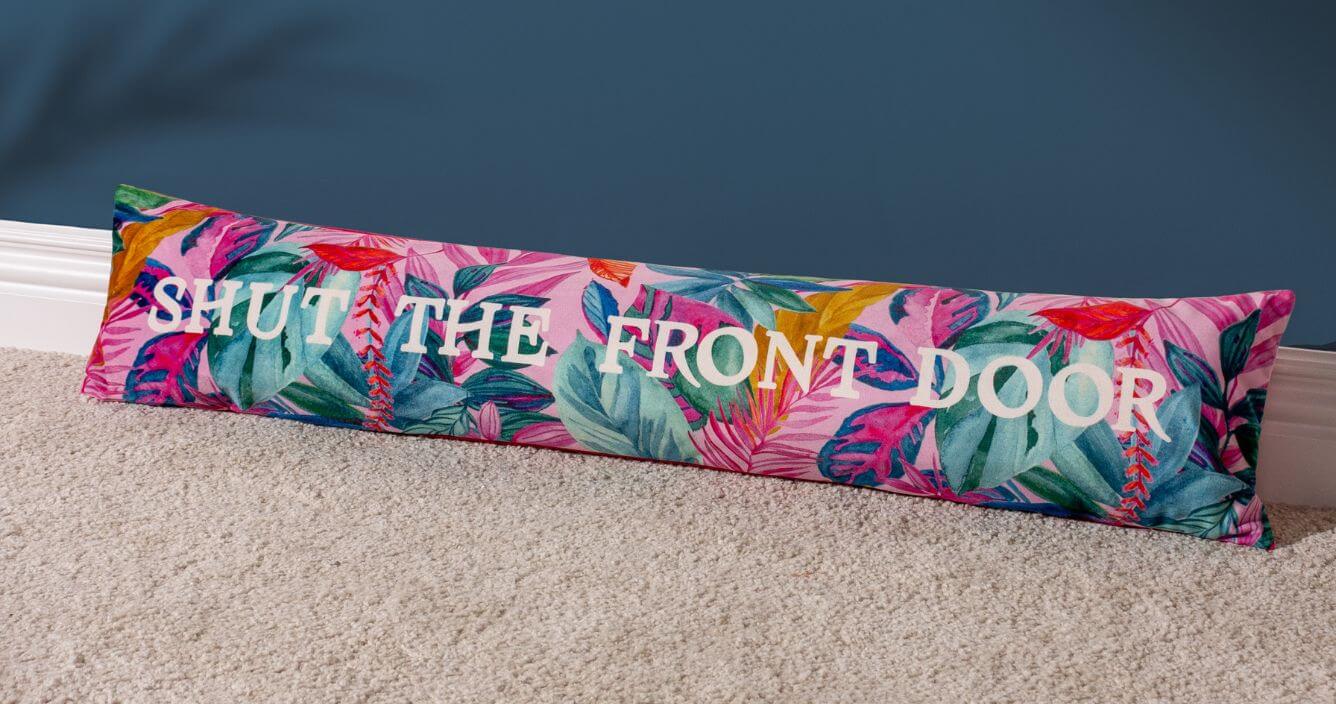 A vibrant draught excluder cushion with a multicoloured exotic leaf design and a printed slogan reading 'shut the front door', laid on a grey carpet in front of a dark blue wall.