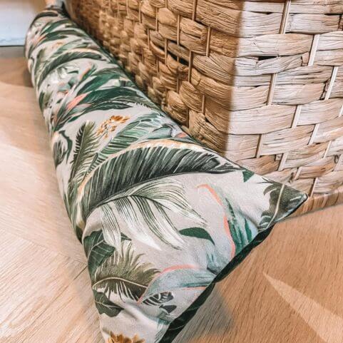 A forest green draught excluder with a printed rainforest leaf design, laid on a wooden floor against a woven storage box.