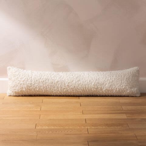 A white draught excluder with an all-over textured bouclé design, laid on a wood panelled floor in front of a neutral wall.