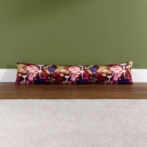A burgundy draught excluder with a printed fungi design, laid behind a neutral rug on a dark wood floor in front of an earthy green wall.