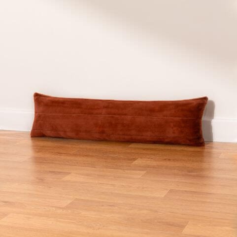 A rusty orange coloured draught excluder cushion with a plain faux fur design, laid on a wooden floor in front of a neutral wall.