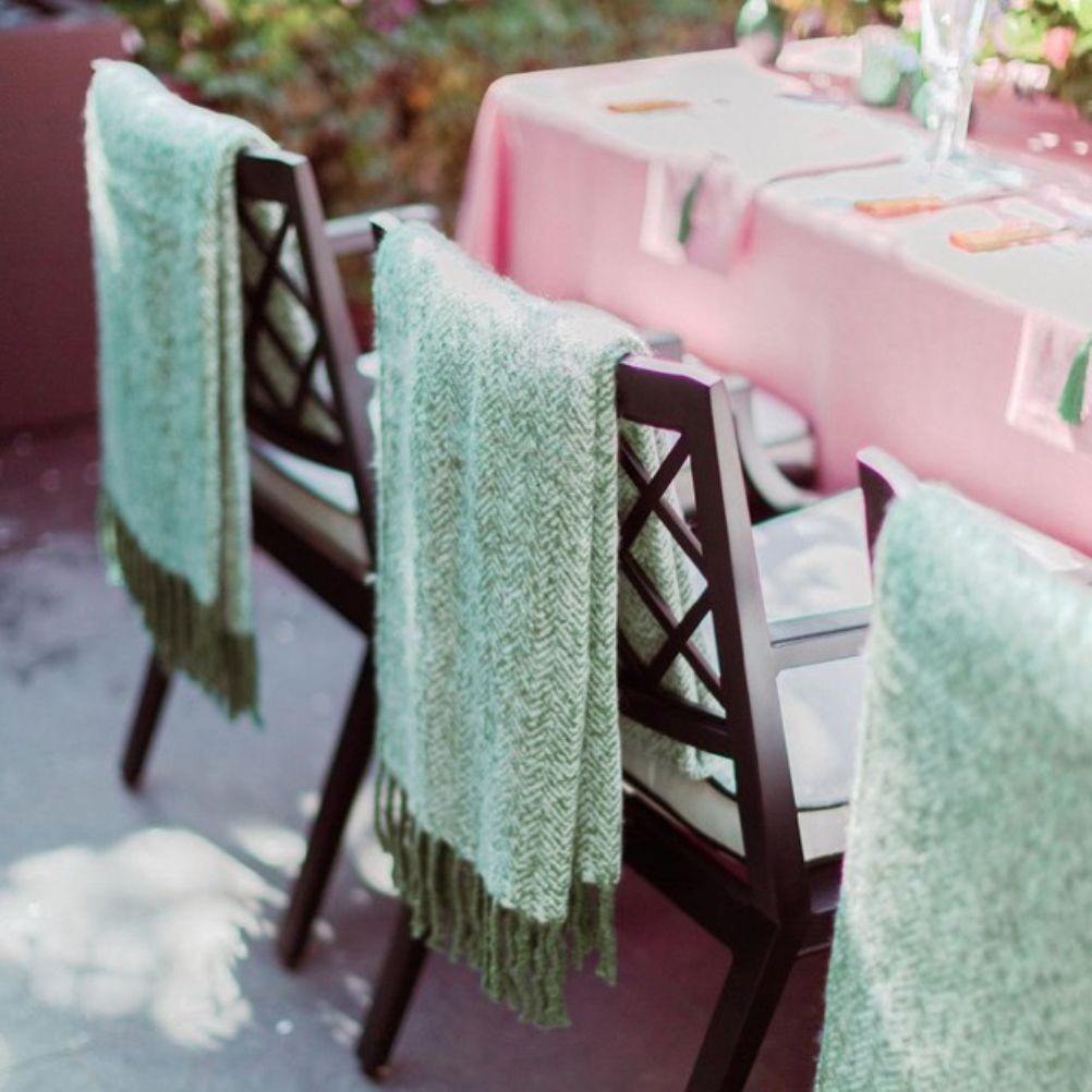 Green fringed throws over the back of dining chairs. The table cloth is pink and set with flowers. There's greenery in the background.