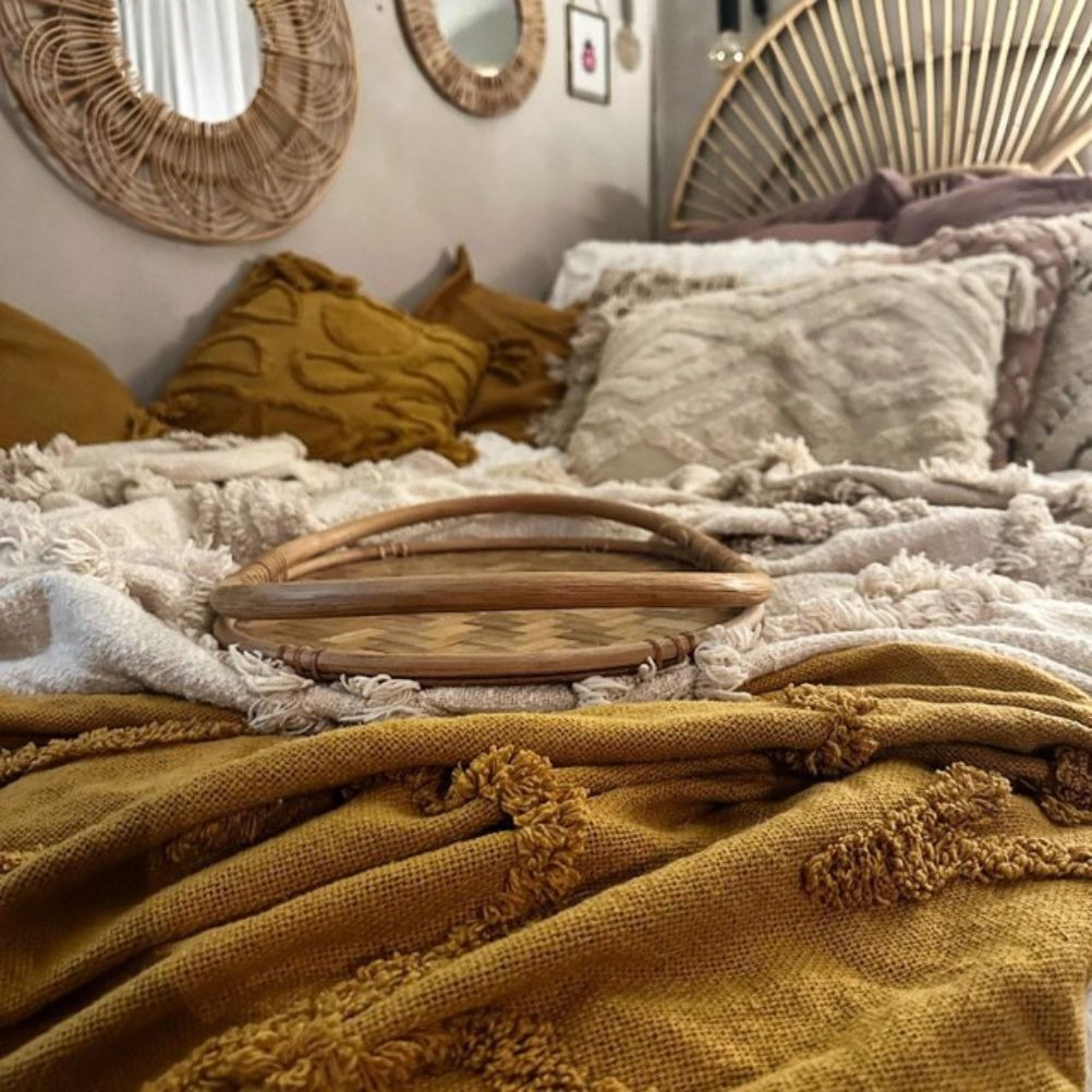 A boho bed layered up with tufted throws and tufted cushions all in cream and ochre. There's a rattan tray in the middle. 