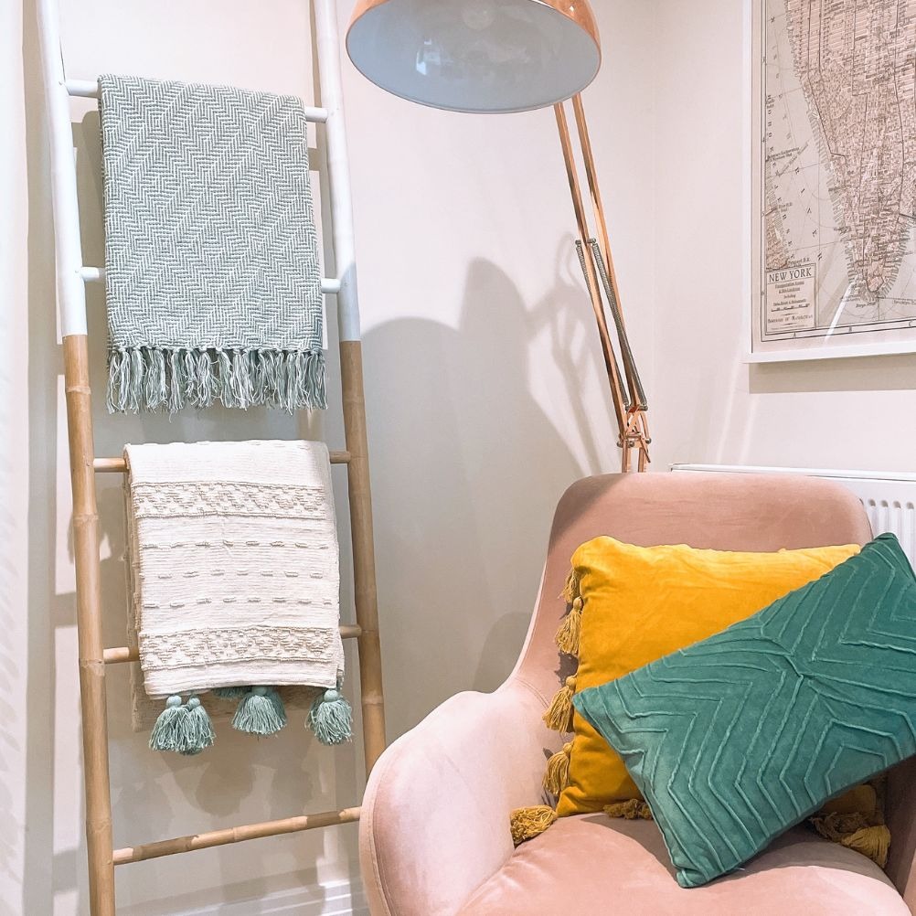 A corner of a living room. There's a ladder that has two fringed throws neatly folded over it. One is seafoam green and the other is cream. The ladder is next to a pink velvet chair with yellow and green velvet cushions on it. Behind is an anglepoise lamp.