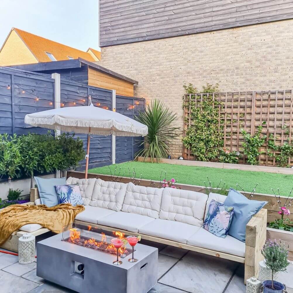 An garden scene with a large outdoor sofa in the foreground. It's layered with a gold fleece throw and lots of cushions. There's a lit firepit in front of the sofa and a parasol behind it.  