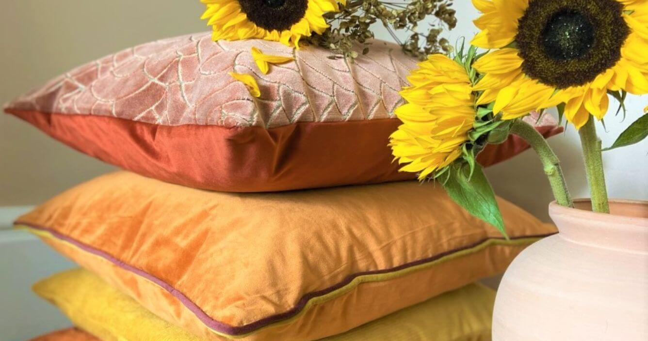 A selection of bright velvet scatter cushions in yellow, orange and red shades, stacked in a single pile behind a vase of sunflowers.