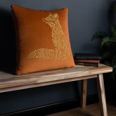 A rust and gold coloured velvet cushion with an embroidered woodland design of a fox, resting on a wooden bench in front of a blue background.