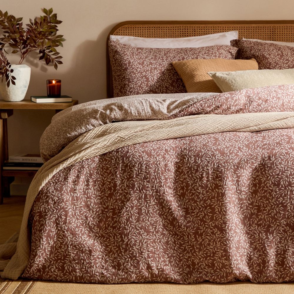 A maroon duvet cover set with a small leaf print in cream, with an orange and cream cushion and a cotton cream throw draped over the top